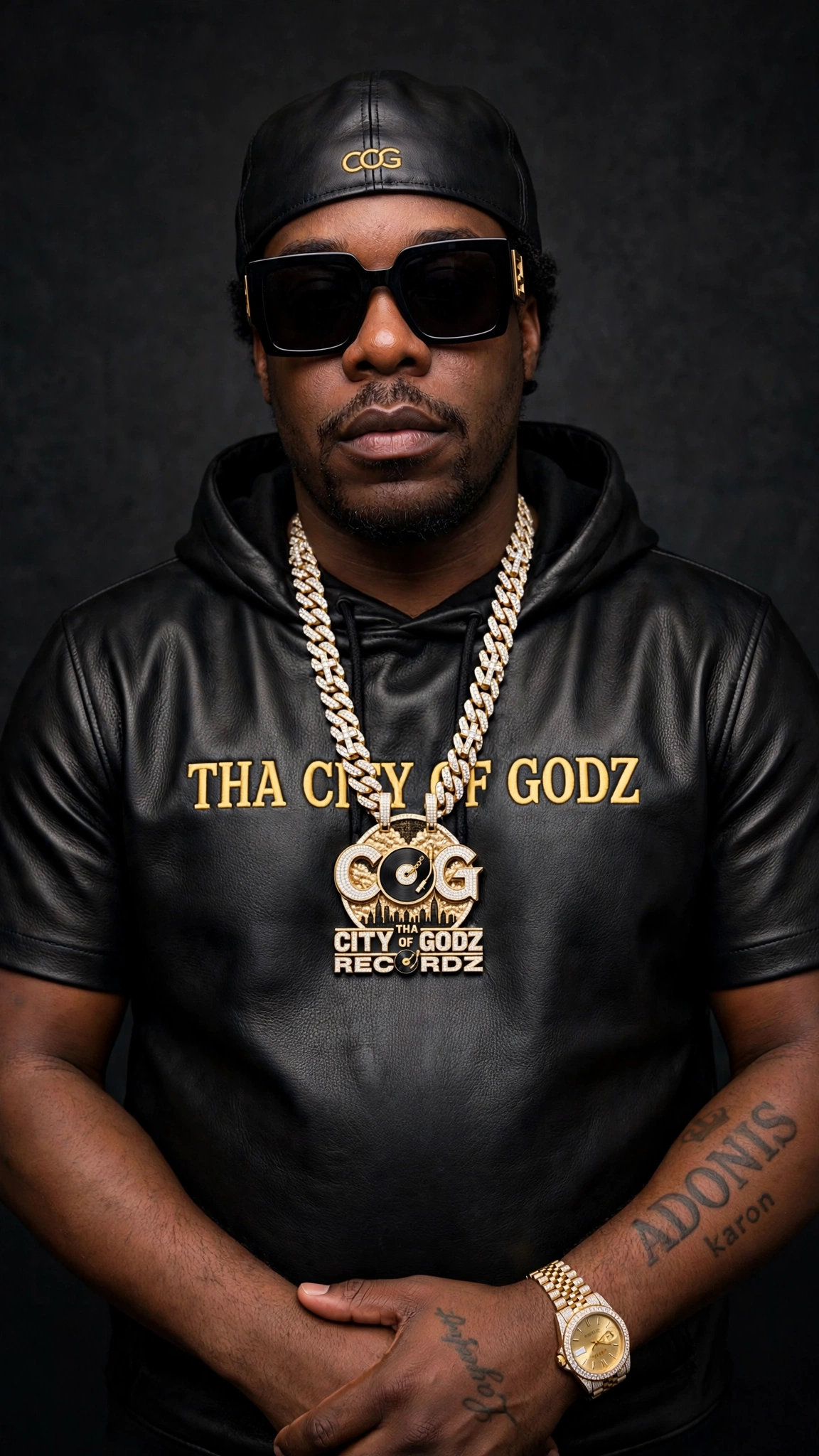 Studio portrait of COG KA'RON in a black leather Tha City of Godz hoodie, COG pendant, black cap and sunglasses