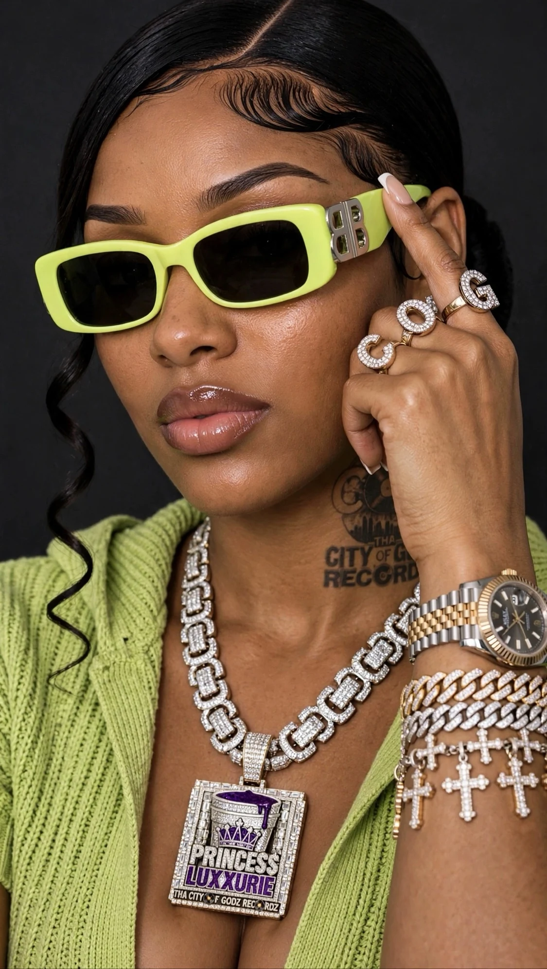 PRINCE$$ LUXXURIE studio portrait in a lime-green Balenciaga hoodie and sunglasses, wearing her iced PRINCESS LUXXURIE drank-cup pendant, COG letter rings and bust-down watch